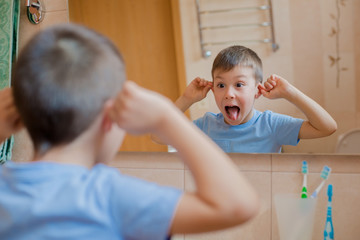 The child grimaces before the mirror in the bathroom. A little boy shows his tongue and has fun.