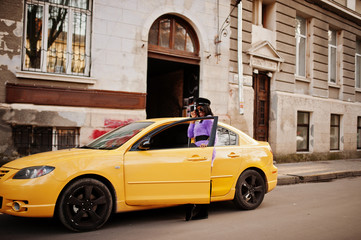 African american woman at violet dress and cap posed at yellow car.