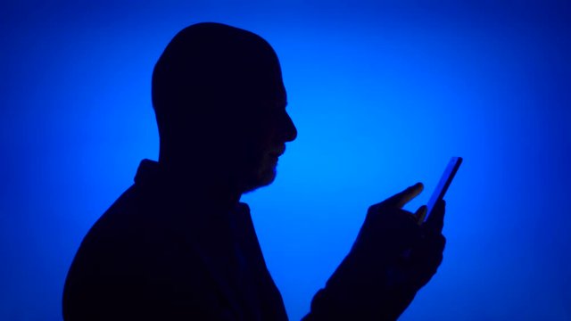 Silhouette of senior man using mobile on blue background. Male's face in profile browsing, reading, chatting online with family and friends on cellphone. Black contur shadow of grandfather's half-face