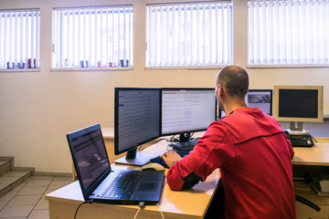 The engineer monitors the computer network. The system administrator looks at multiple monitors. A man is sitting at the office behind a worker's desk.