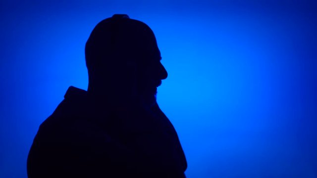 Silhouette of senior man putting on big black wireless headphones on blue background. Male's face in profile listening to music. Black contur shadow of grandfather's half-face singing