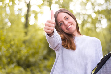 Caucasian european young woman or girl university or student, entrepreneur with positive attitude thumbs up, affirmative gesture, in nature with erasmus folder, study trip learned languages