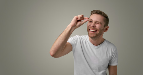 The Jolly young man has bristles, giggles at camera and shows off something small, dressed casually, isolated on a white background. Happy Caucasian shows size refers to something very tiny or small - obrazy, fototapety, plakaty