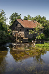 Obraz premium Ancient abandoned water mill surrounded by beautiful nature. House built of stone and wood, exterior walls and dilapidated bridge on river is reflection of trees and hous ruins