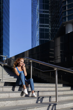 Beautiful girl sitting on the steps with her smartphone between skyscrapers