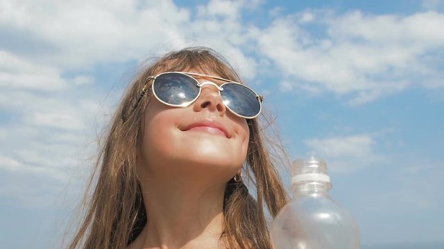The child on vacation drinks water. Little girl in sunglasses drinks water against the background of the sky.