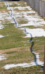 Grass showing through between clumps of melting snow behind a chain link fence.