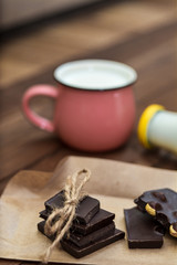 Handmade chocolate pieces on rustic paper, a cup and a bottle with milk on a wooden background. Delicious breakfast - milk and chocolate.