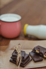 Handmade chocolate pieces on crafting paper, a cup and a bottle with milk on a wooden background. Delicious breakfast - milk and chocolate.