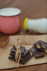 Handmade chocolate pieces on crafting paper, a cup and a bottle with milk on a wooden background.