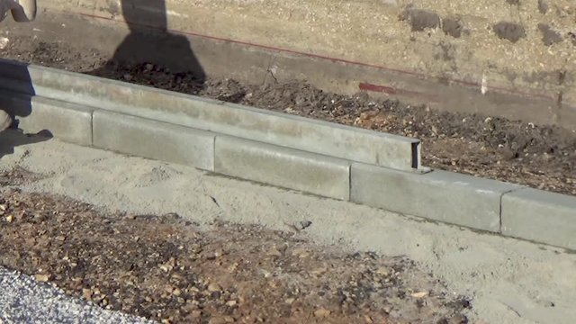 worker installing concrete curb stone and using string with metal stakes to level at sidewalk construction site.