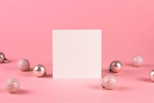 Mock up with square invitation card on trendy pastel light pink background with christmas ornaments. Greeting card and silver Christmas baubles. Minimal christmas concept.