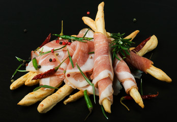 Grissini bread sticks with prosciutto on a black background with rosemary, pepper 