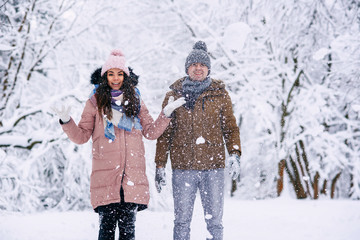 A beautiful young woman and man in warm clothes rejoices at falling snow in a winter park.