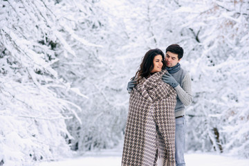 Fototapeta na wymiar Happy lovely couple covered with a warm blanket and walking in a snowy park. Pregnancy concept.