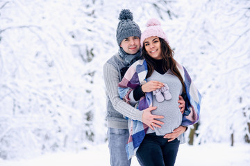 Man holding woman's pregnant belly and the woman holding baby shoes in her hand while they stand on snowy winter park.