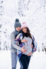 Fototapeta na wymiar Man holding woman's pregnant belly and the woman holding baby shoes in her hand while they stand on snowy winter park.