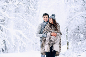 Fototapeta na wymiar Young man hugs tenderly his beautiful pregnant wife covered in warm blanket with snowfall on background. Family love and care concept.
