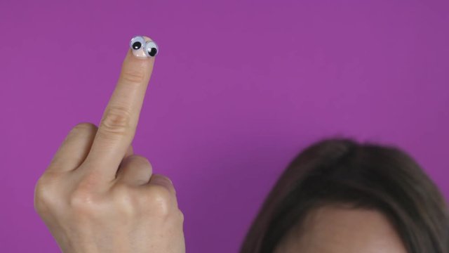 Female womans hand with stick on googly eyes. Giving the finger. Her head defocused in the background.