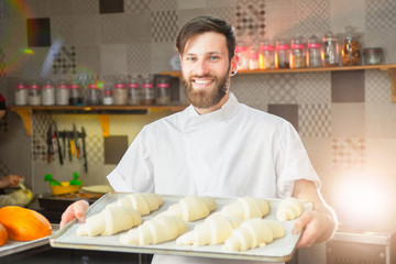 A young handsome baker is holding raw croissants of white dough in the hands of an oven. Semi-finished products from dough