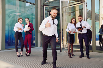 A young male businessman is enjoying a successful deal on the background of office workers. Demonstration of positive emotions and gestures. Career. Success. Shows a thumbs-up.