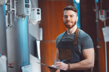 A young handsome brewer in an apron with a tablet in his hands amid the vats of a brewery