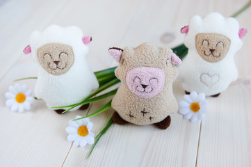 three soft toy sheep handmade on a light wooden background, artificial flowers, grass