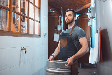 A young brewer in an apron holds a barrel with beer in the hands of a brewery - Powered by Adobe