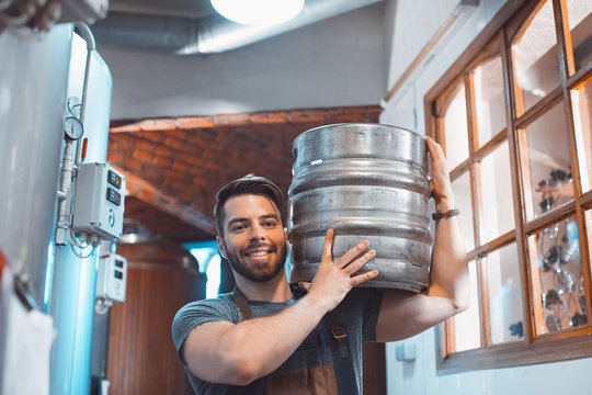 A young brewer in an apron holds a barrel with beer in the hands of a brewery