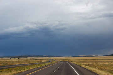 The desert highway among the steppe, in front of the blue mountains. Summer day, dark sky, big dark blue clouds in the sky