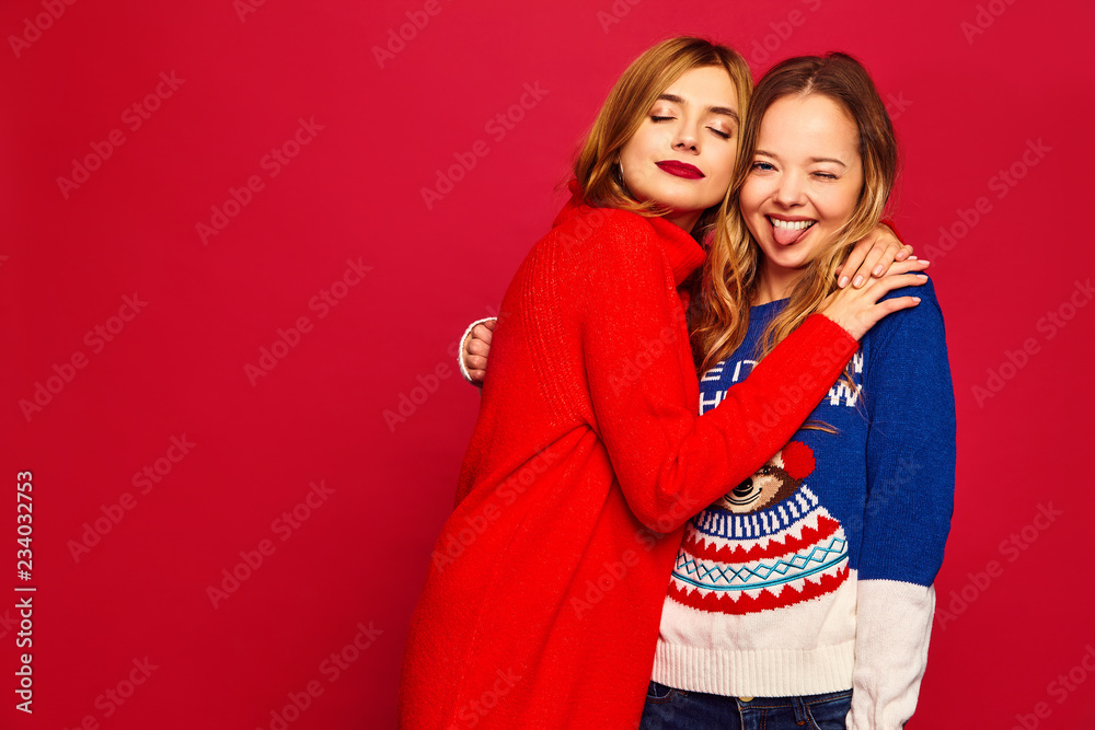 Wall mural Two beautiful smiling gorgeous girls looking at camera. Women standing in stylish winter warm sweaters on red background. Christmas, x-mas, concept.