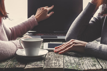 Teamwork,two young business women sitting at table,drinking coffee and working online.First girl is showing pen on chart and diagram on computer screen.On table tablet PC.Students are learning online.