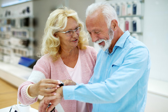Senior couple choosing new smart watch. Man putting watch on hand while his wife helping him. Tech store interior.