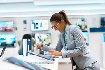 Happy Caucasian woman with ponytail dressed in business clothes looking fort new tablet to by while standing near stand in tech store.
