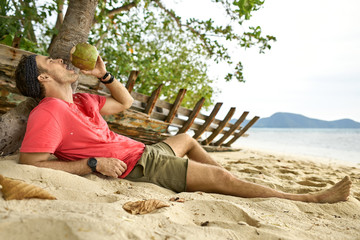 Man with stubble is drinking from coconut on sand beach