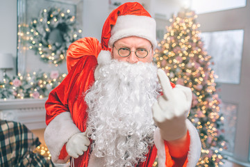 Rude and angry man wears Santa Claus clothes. He shows fuck on camera. Guy holds red bag with one hand.
