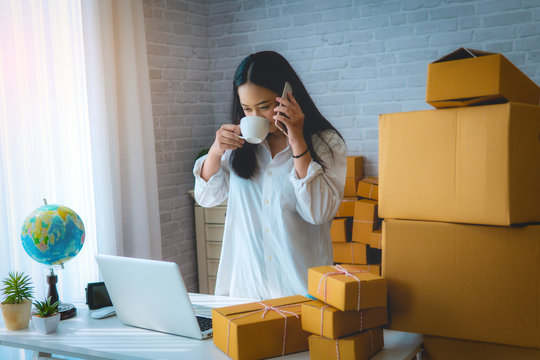 Young Women Holding A White Coffee Mug Small Business Owner Working At Home Office Packaging On Background. Online Shopping SME Entrepreneur Or Freelance Working Concept.