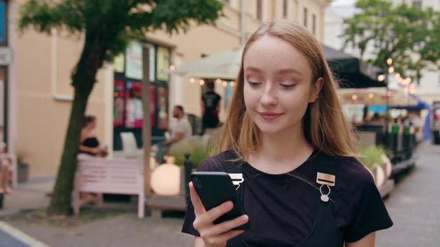An attractive young red-haired lady using a phone in town. Close-up dolly shot. Soft focus