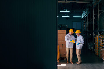 Fototapeta premium Two Caucasian workers discussing while younger worker holding documents. Warehouse interior.