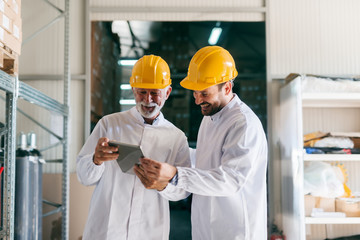 Senior worker showing to coworker documents on tablet. Warehouse interior.
