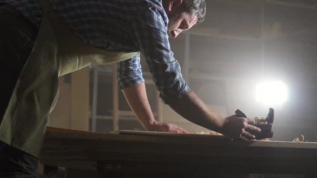 male carpenter in shirt in dark workshop planing wood
