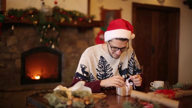 Happy man wearing santa hats opening Christmas gift box near fireplace with flashing garland. Smiling guy recieved a parcel with presents and enjoying unpacking package. Winter holidays concept.