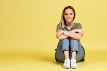 Fototapeta premium Cute young girl smiling with big brown eyes sits on the floor on a yellow background. Place for text