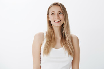 Obraz premium Dreamy girl feeling nostalgic, recalling nice moment from past gazing at upper left corner with delighted happy smile posing carefree against gray background in casual white tank-top