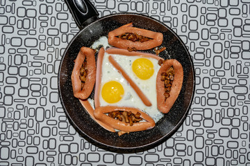 Cast-iron frying pan with fried eggs, sausage, mushroom and beans on wooden table. Top view.