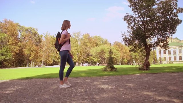 side view student with backpack. girl with blond hair wearing pink t-shirt and jeans stroll in park on the background beautiful old college