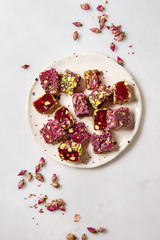 Variety of traditional turkish dessert Turkish Delight different taste and colors with rose petals and pistachio nuts on ceramic plate over white marble background. Flat lay, space