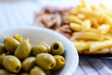 Green olives in white bowl on kitchen table with meat background and french fries. Background with pork and potatoes on the kitchen table with green olives.