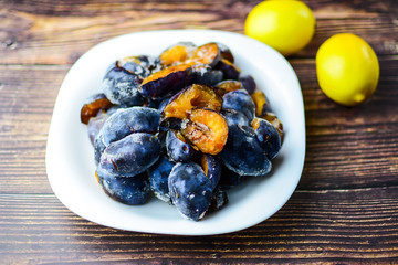 Healthy food concept. Frozen organic plums on a white plate with two lemons on a rustic wooden table. 