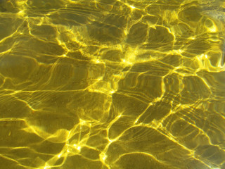 Sunlight refracting through transparent water and creates sun glare on golden sand of bottom. Top view of yellow ripple texture of liquid surface and sunny shining reflections.